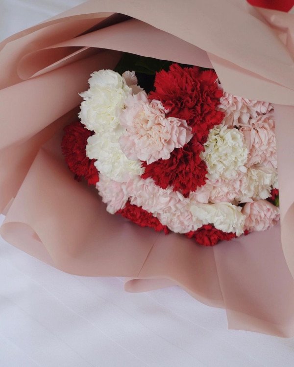Bouquet of red, white, and pink carnations wrapped in pink paper on a white surface.