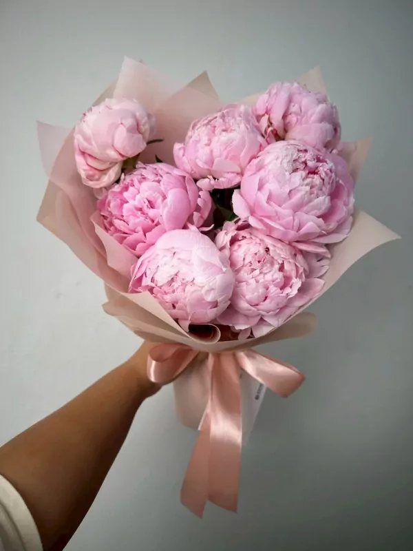 A hand holds a bouquet of seven pink peonies wrapped in light pink paper and tied with a satin ribbon.