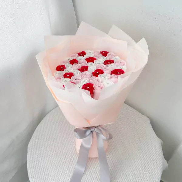 Bouquet of pink, red, and white roses with diamond accents, wrapped in sheer pink paper and tied with a grey ribbon.