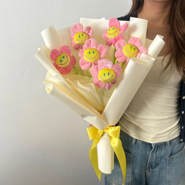 Pink plush smiley face daisies with yellow centers in a bouquet wrapped in pale yellow and cream paper, tied with a yellow ribbon.