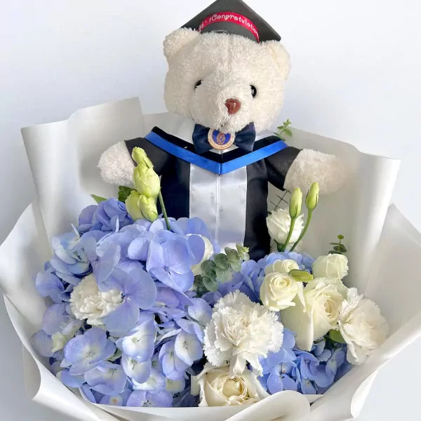 Teddy bear in graduation gown holding a bouquet of blue hydrangeas and white roses.