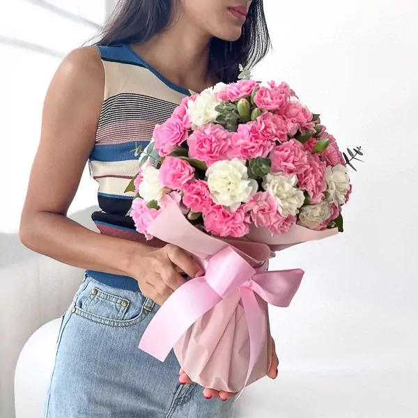 Woman in striped top and jeans holding pink and white carnation bouquet wrapped in pink paper with a pink bow.