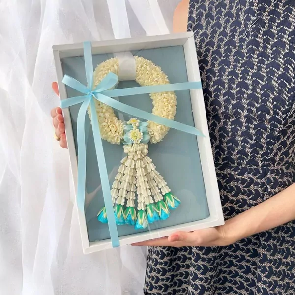 White jasmine malai with blue ribbon in a white box, held by a person in a navy patterned dress.