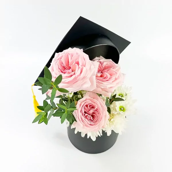 Pink roses and white mums in a black graduation hat box with a yellow tassel.
