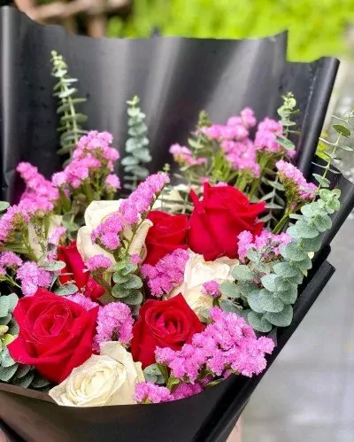 A bouquet of red and white roses with pink statice flowers, wrapped in black paper.