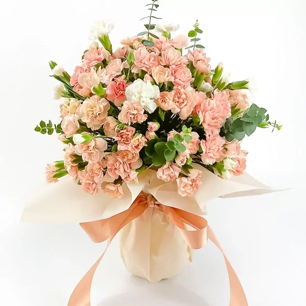 A bouquet of peach and white carnations with eucalyptus, wrapped in pale orange paper and tied with a peach ribbon.