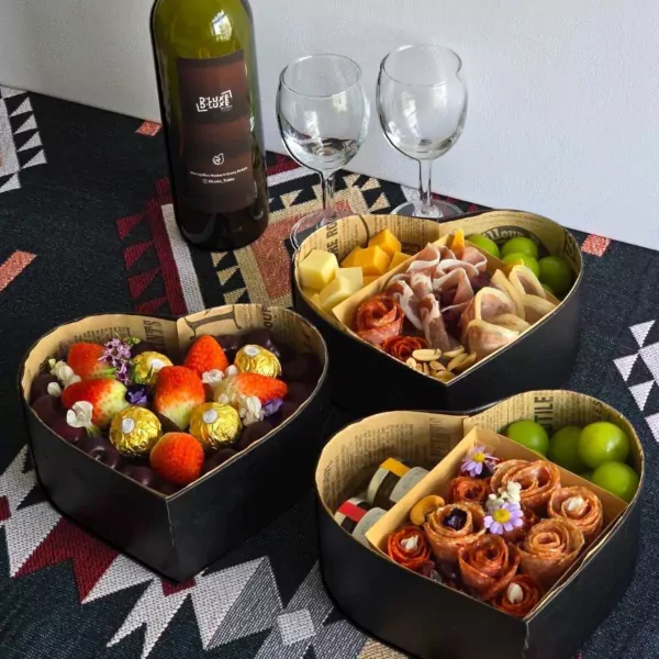 Heart-shaped charcuterie box with grapes, cheese, cured meats, and strawberries, next to wine and glasses.