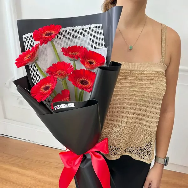 Red gerbera daisy bouquet wrapped in black paper, tied with a red ribbon, held by a person wearing a tan top.