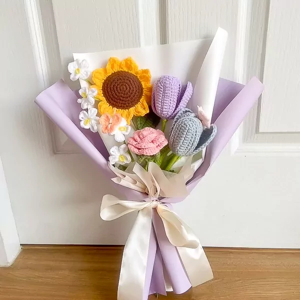 Crocheted bouquet with sunflower, tulips, roses, and white flowers wrapped in purple paper, tied with a ribbon.