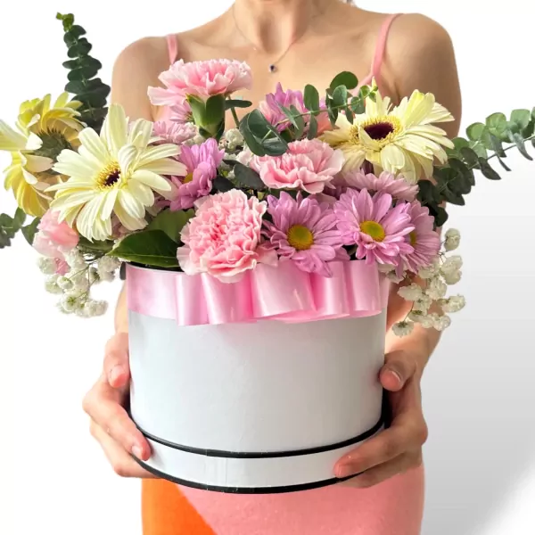A woman holds a round white flower box adorned with a pink ribbon, filled with pink and yellow gerbera daisies, carnations, and eucalyptus.