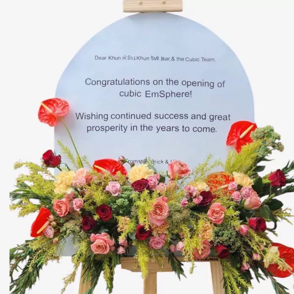 Floral stand with red anthuriums, pink roses, and greenery on a congratulatory sign.