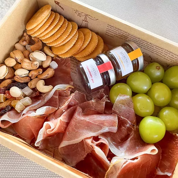 Charcuterie box with Serrano ham, crackers, nuts, fruit, and two small jars of jam and honey.