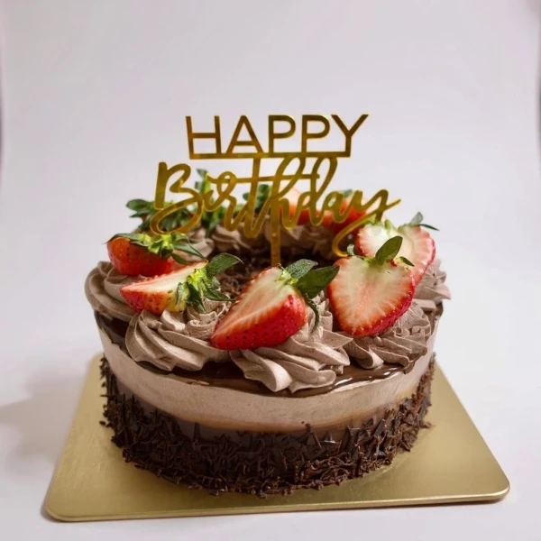 Chocolate cake with chocolate frosting, fresh strawberries, and a "Happy Birthday" topper on a gold cake board.