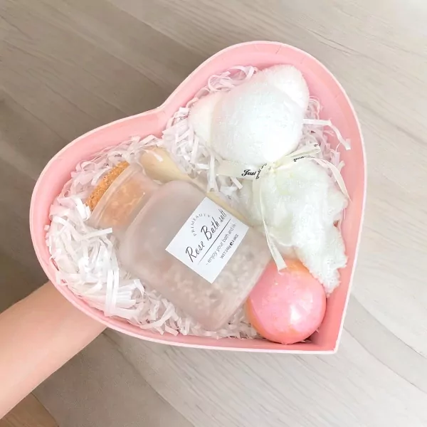Heart-shaped pink gift box with a teddy bear towel, rose bath salts, bath bomb, and wooden spoon.
