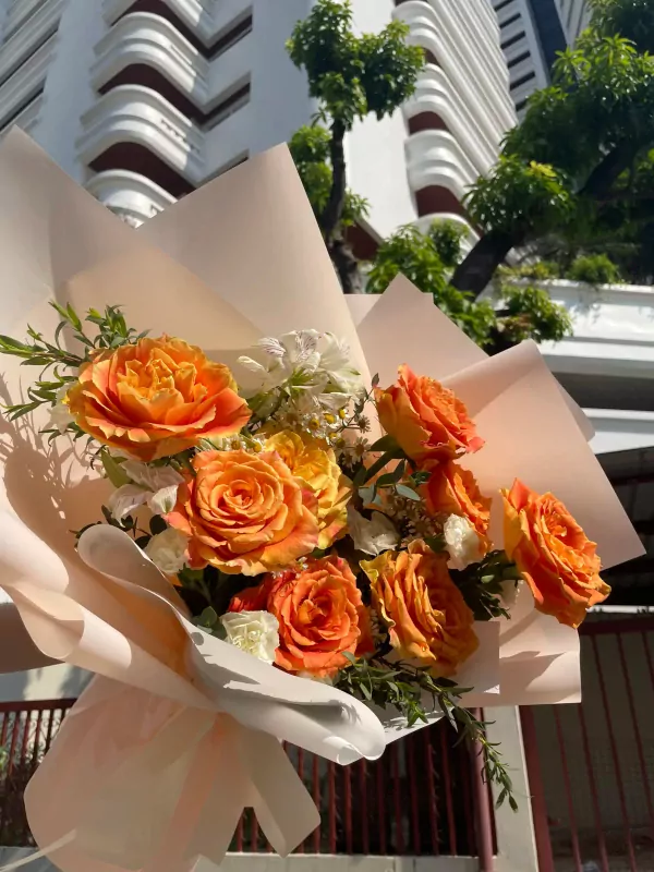Bouquet of vibrant orange roses, alstromerias, and daisies wrapped in pale pink paper.