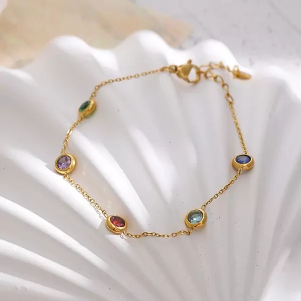 Gold chain bracelet with colorful gemstones, including purple, green, red, blue, and teal, on a white, wavy surface.