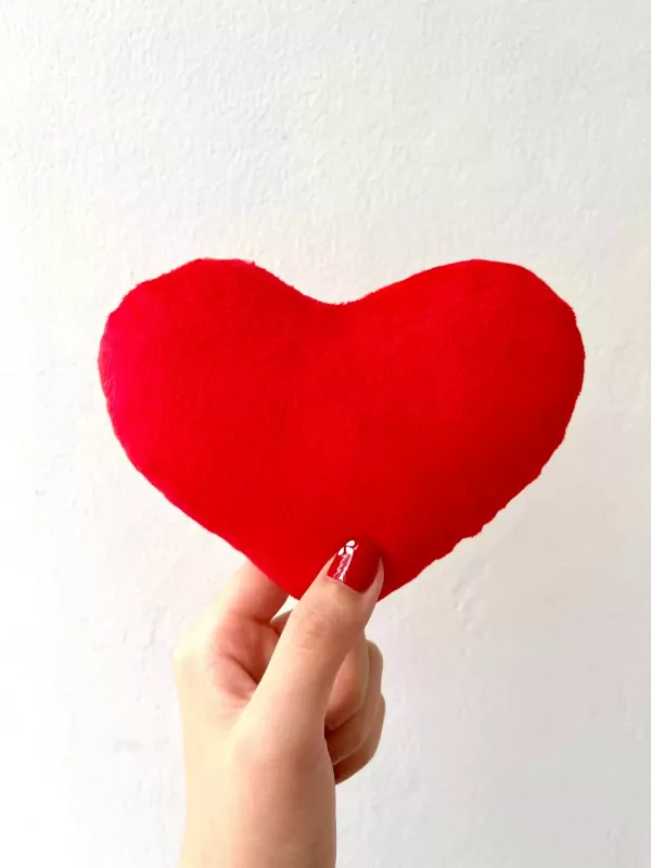 Hand holding a red heart pillow against a white wall, with a heart design on red nail polish.