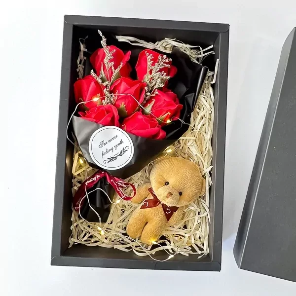 A gift box with red soap roses, a teddy bear keychain, and fairy lights.
