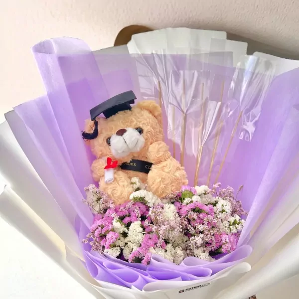 Graduation teddy bear in a bouquet of purple and white dried flowers, wearing a graduation cap.