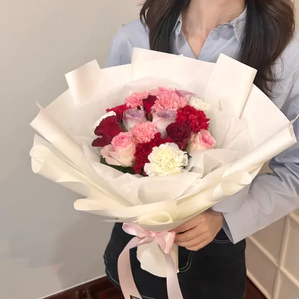 Woman holding a bouquet of pink, red, and white roses and carnations, wrapped in white paper with a pink ribbon.