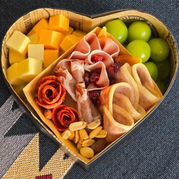 Heart-shaped box filled with cheese cubes, charcuterie, green grapes, and nuts.