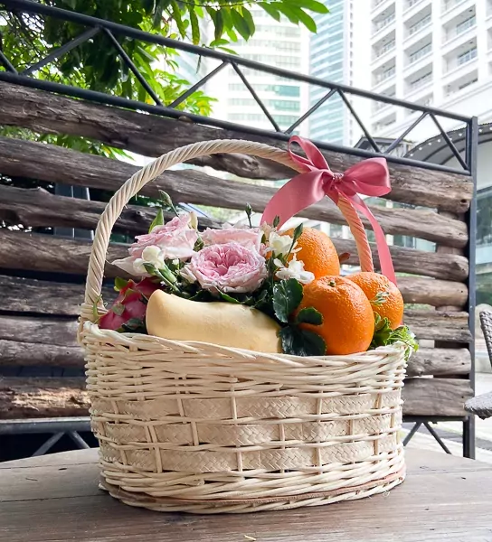 A woven basket filled with pink roses, mango, dragon fruit, and oranges, tied with a pink ribbon.