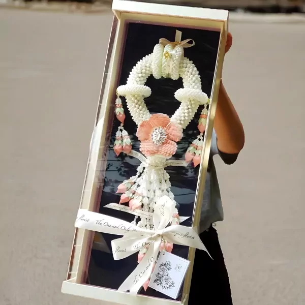 A person holds a gift box containing a large, white jasmine garland with a pink flower centerpiece.