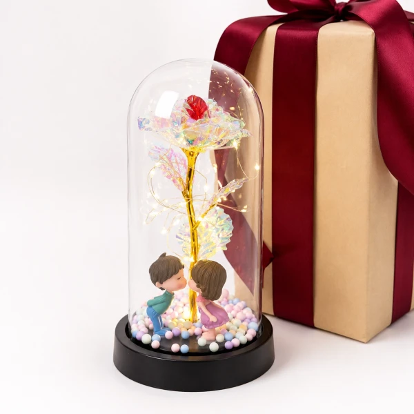 Glass dome with holographic rose, kissing couple figurines, and fairy lights next to a wrapped gift.