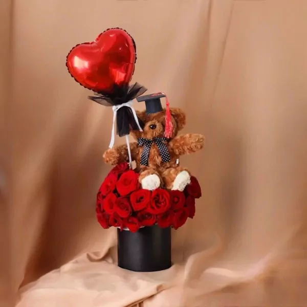 Graduation teddy bear wearing cap, holding heart balloon, surrounded by red roses in a black box.