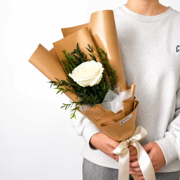 Hand holding a white rose bouquet with eucalyptus, wrapped in brown paper and tied with a white ribbon, against buildings.