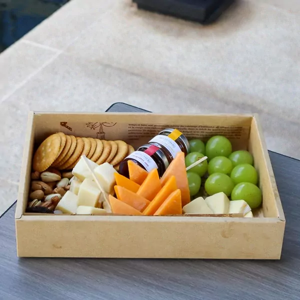 Cheese platter with crackers, nuts, grapes, cheese slices, and small jam jars in a wooden box.