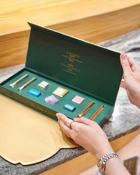 Green box of assorted chocolates, including bars and jewel-shaped pieces, held by a person with a ring and watch.