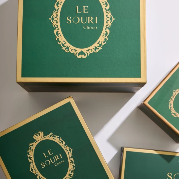 Green Le Souri chocolate boxes with gold trim and ornate emblem, presented on a white surface.