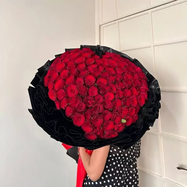99 red roses arranged in a heart shape, a large bouquet with black wrapping and a red bow.