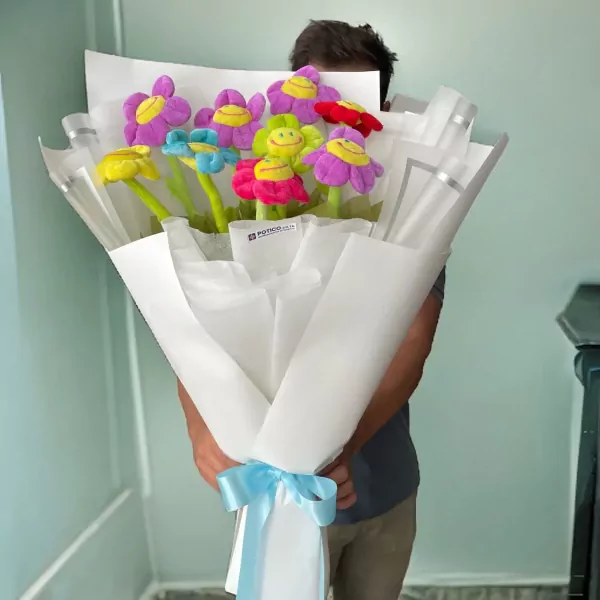 Bouquet of colorful plush smiley-face daisies in white and silver wrapping, tied with a light blue ribbon.