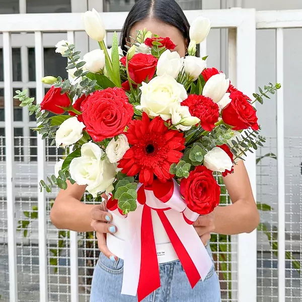 A round white box overflowing with red and white roses, tulips, gerbera, carnations, and eucalyptus, tied with a red and white ribbon.