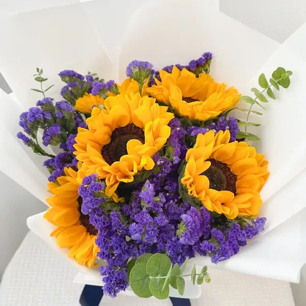 A round bouquet of sunflowers and purple statice flowers wrapped in white paper.