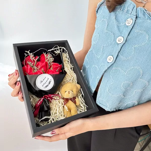 A gift box filled with red soap roses, a teddy bear keychain, and straw.