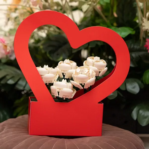 A red heart-shaped basket holding a bouquet of 5 white Lego roses, set against a backdrop of green leaves and pink flowers.