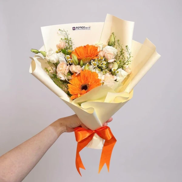 Hand holds a bouquet of orange gerbera daisies, white roses, and delicate green foliage, tied with an orange ribbon.