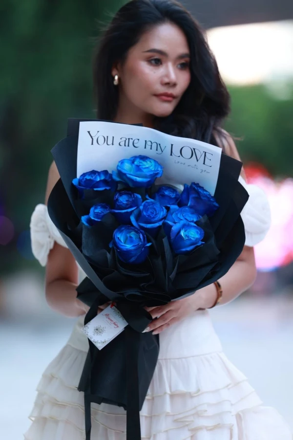 Woman holding a bouquet of vibrant blue roses wrapped in black paper with a card that says "You are my LOVE".