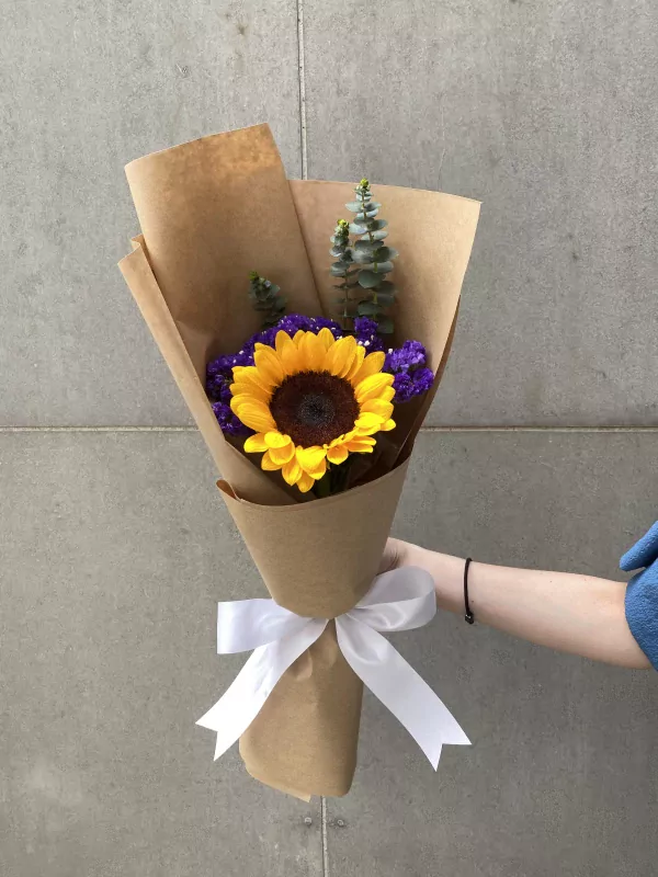 Sunflower bouquet with purple statice flowers, eucalyptus, and a white ribbon, wrapped in brown paper.