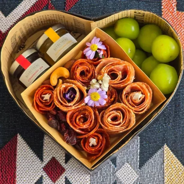 Heart-shaped box with charcuterie roses, jam, nuts, cranberries, green plums, and flowers.