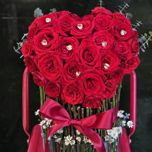 Heart-shaped bouquet of red roses with heart crystals in a clear acrylic cylinder vase.
