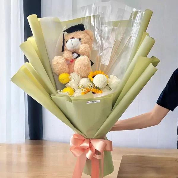 Graduation bouquet with teddy bear, sunflowers, roses, and yellow flowers, wrapped in green and yellow paper, tied with a pink bow.