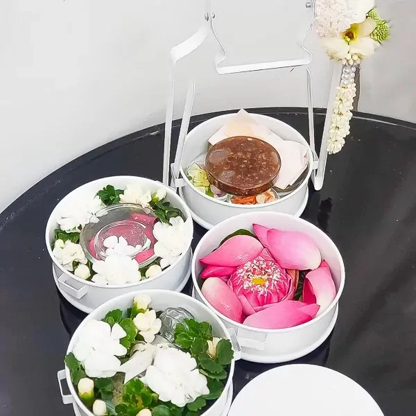 Tiers of white containers holding colorful Mieng Kham ingredients and lotus flowers, arranged in a stand.