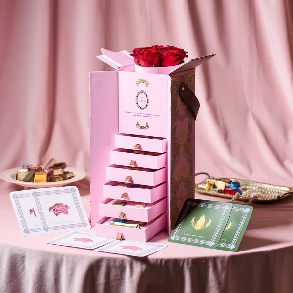 Pink chocolate box with stacked drawers, red roses, chocolates, and cards.