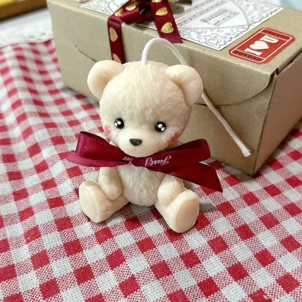 Cream teddy bear candle with red ribbon sits on red and white checkered cloth, with gift box in background.