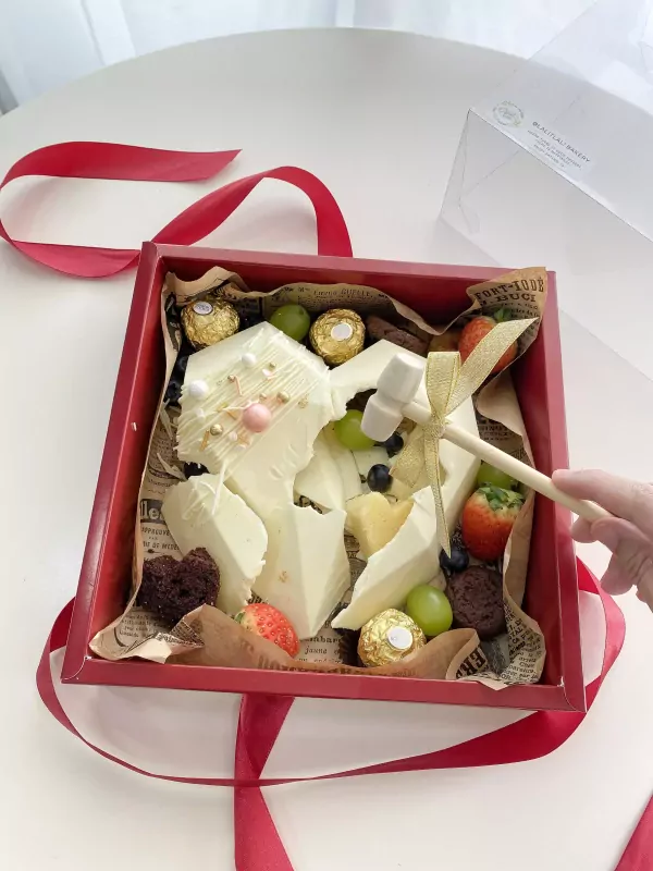 Red box with a white chocolate heart cake, fruits, chocolates, and a hammer.