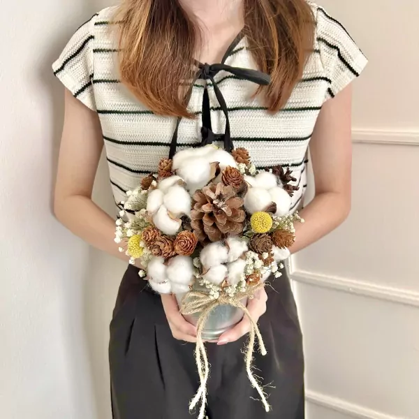 A person holds a rustic bouquet of cotton flowers, pine cones, and yellow accents in a silver pot.